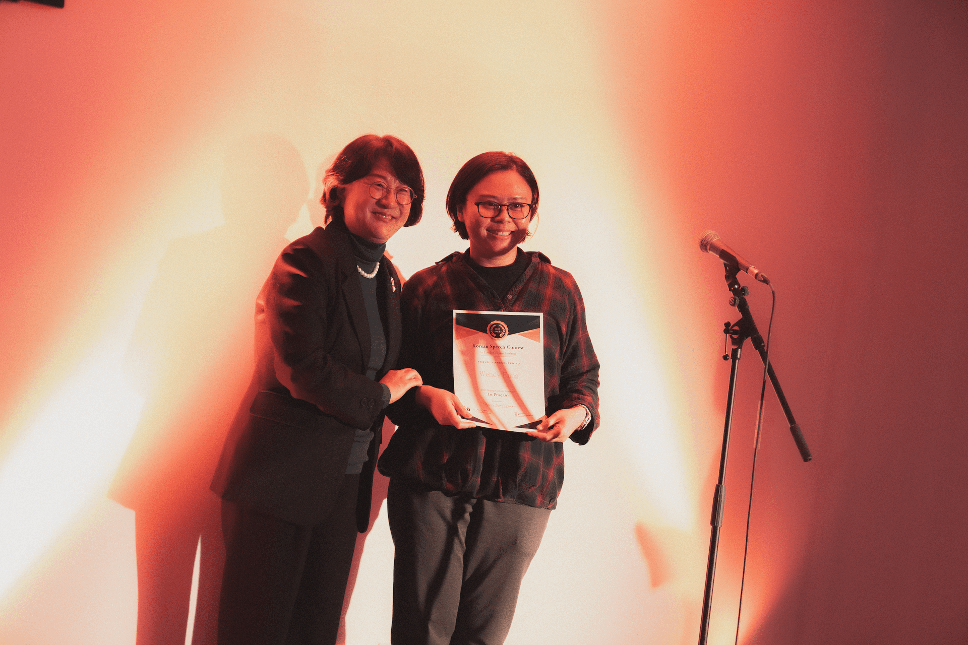 Two persons stand smiling with a certificate under warm lighting, next to a microphone stand.