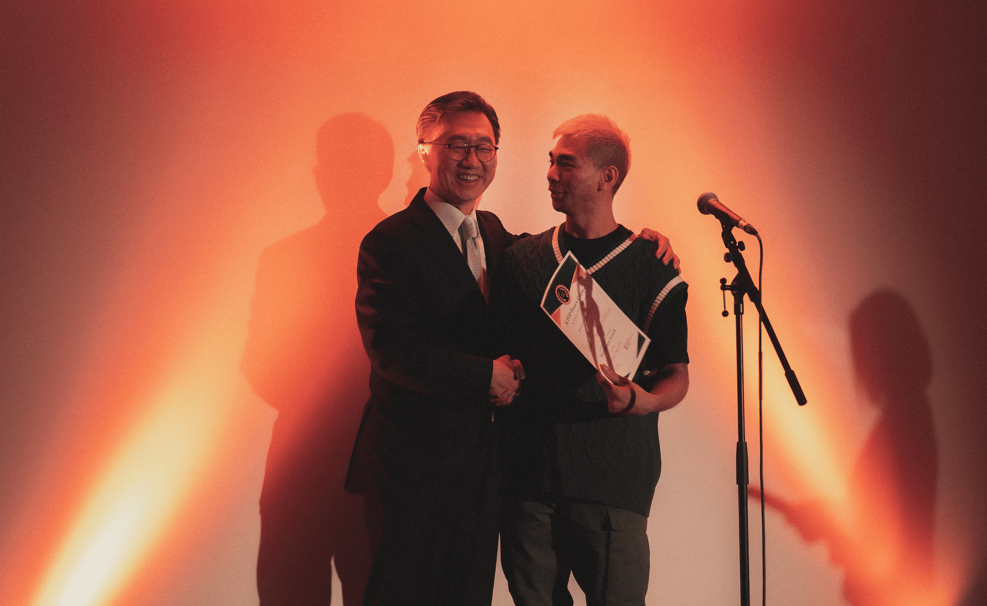 Two people stand together under warm lighting, one holds a certificate, in front of a microphone on a lit stage.