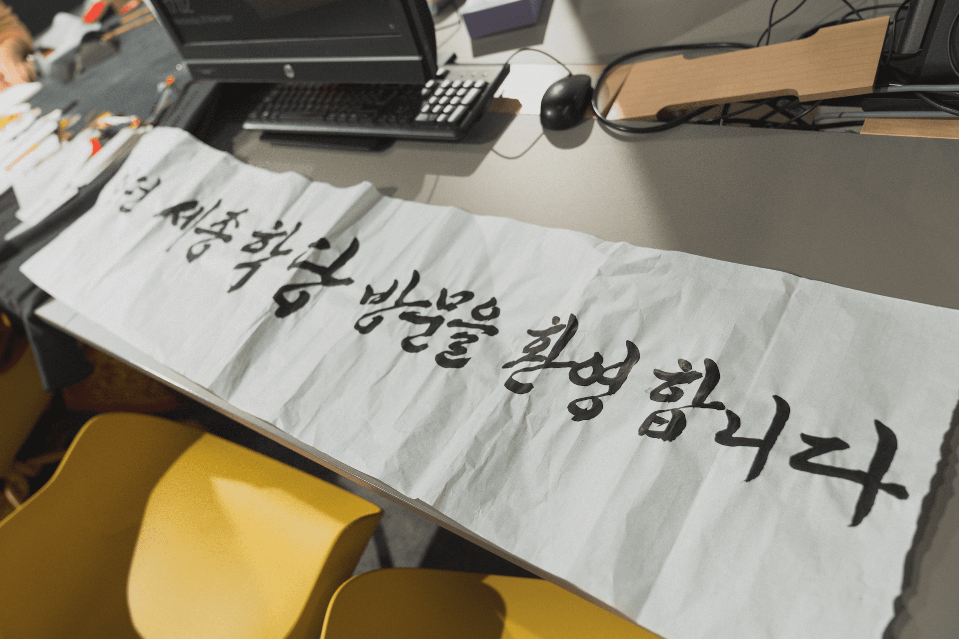 A piece of paper with Korean calligraphy lies on a table near a computer, surrounded by yellow chairs under fluorescent lighting.