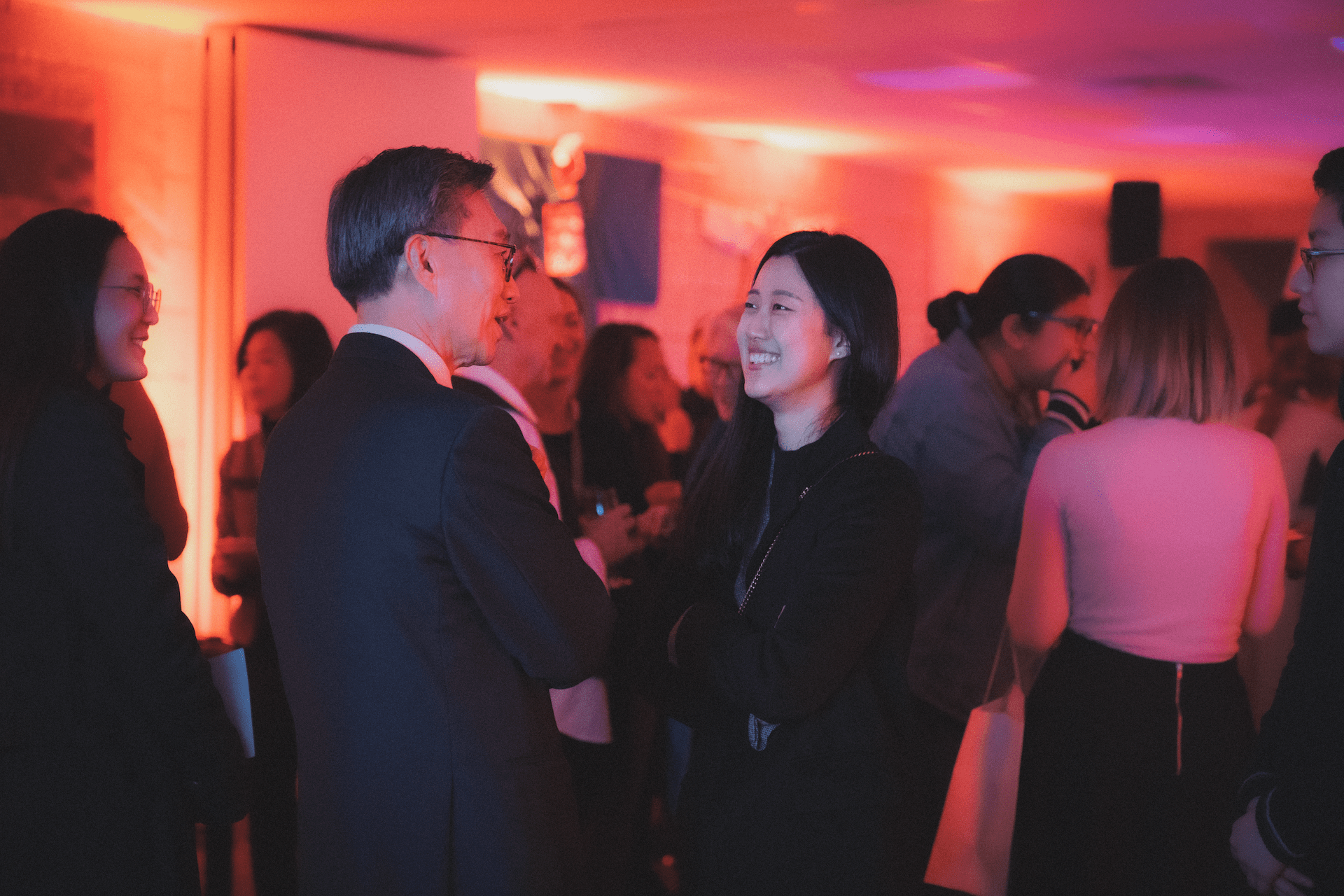 A group of people in formal attire interact at an indoor event with warm lighting and wall decorations.
