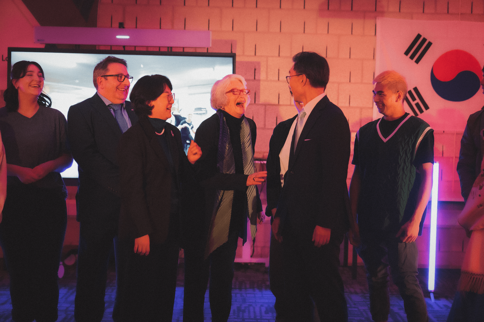 Seven people smiling and interacting indoors near a screen, with a South Korean flag prominently hanging on the wall behind them.