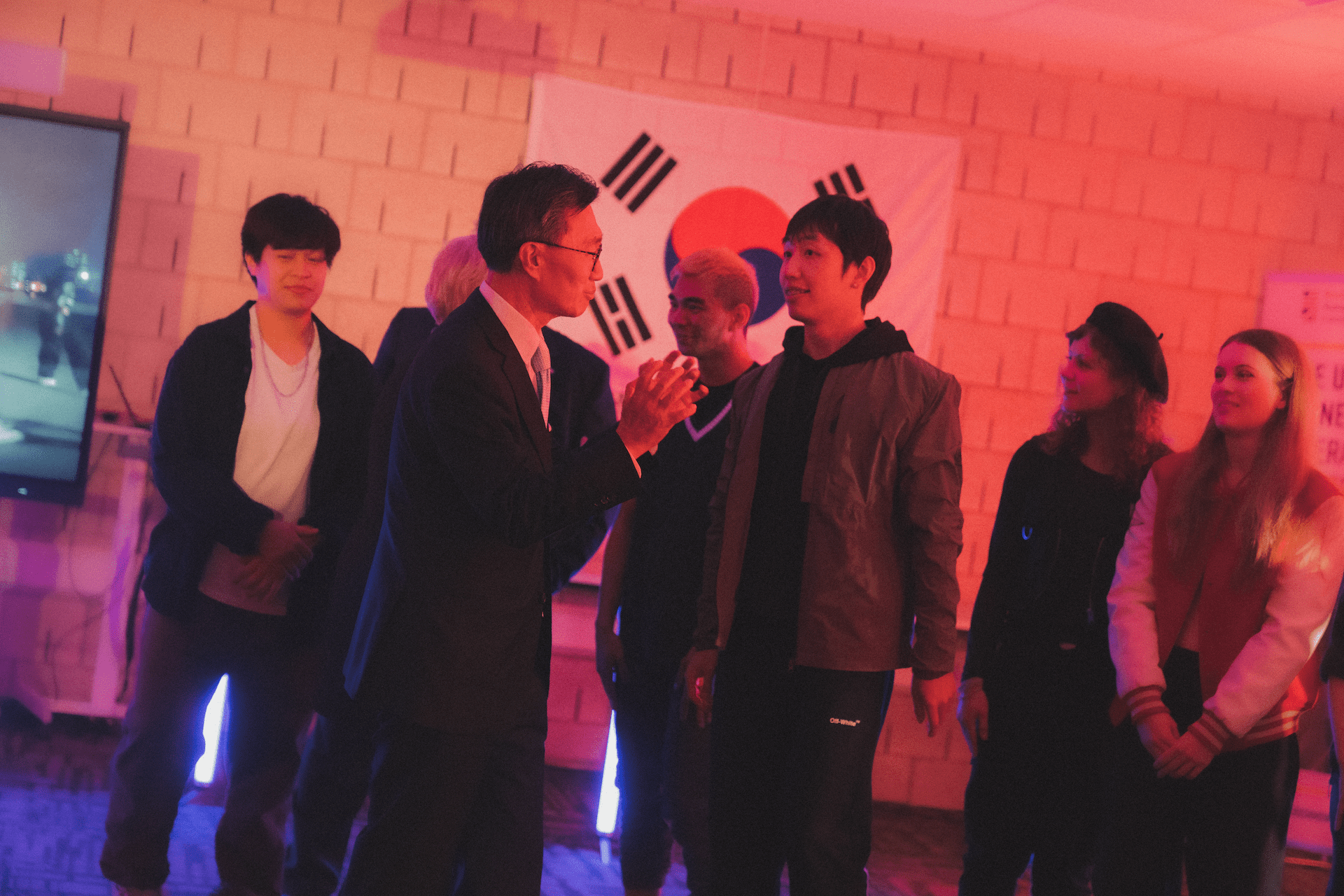 A group of people converse in front of a South Korean flag, illuminated by pinkish-red lighting, creating a lively atmosphere.
