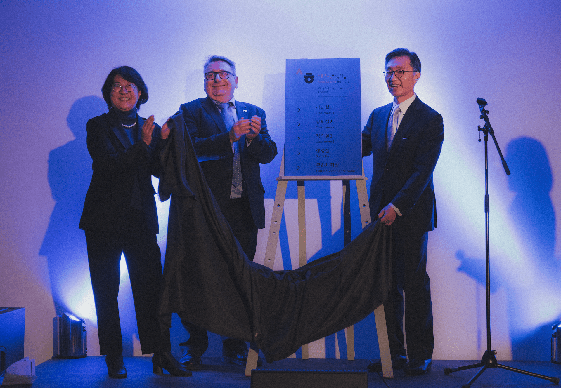 Three people unveiling a plaque on a stage with dramatic blue lighting, and a microphone stand nearby.
