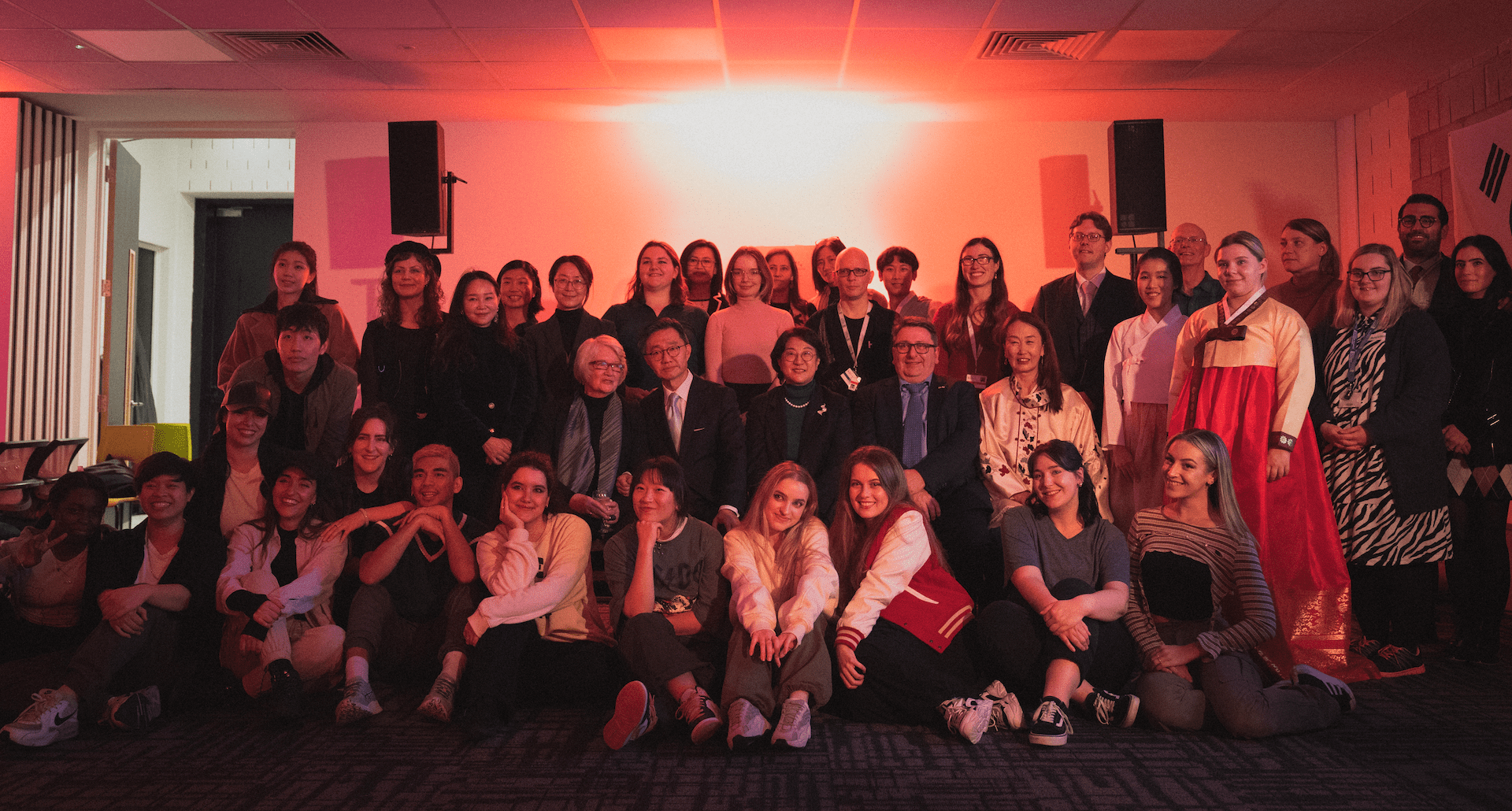 A group of people poses indoors under red lighting. Some are seated, others stand, wearing various styles of clothing, including traditional attire.