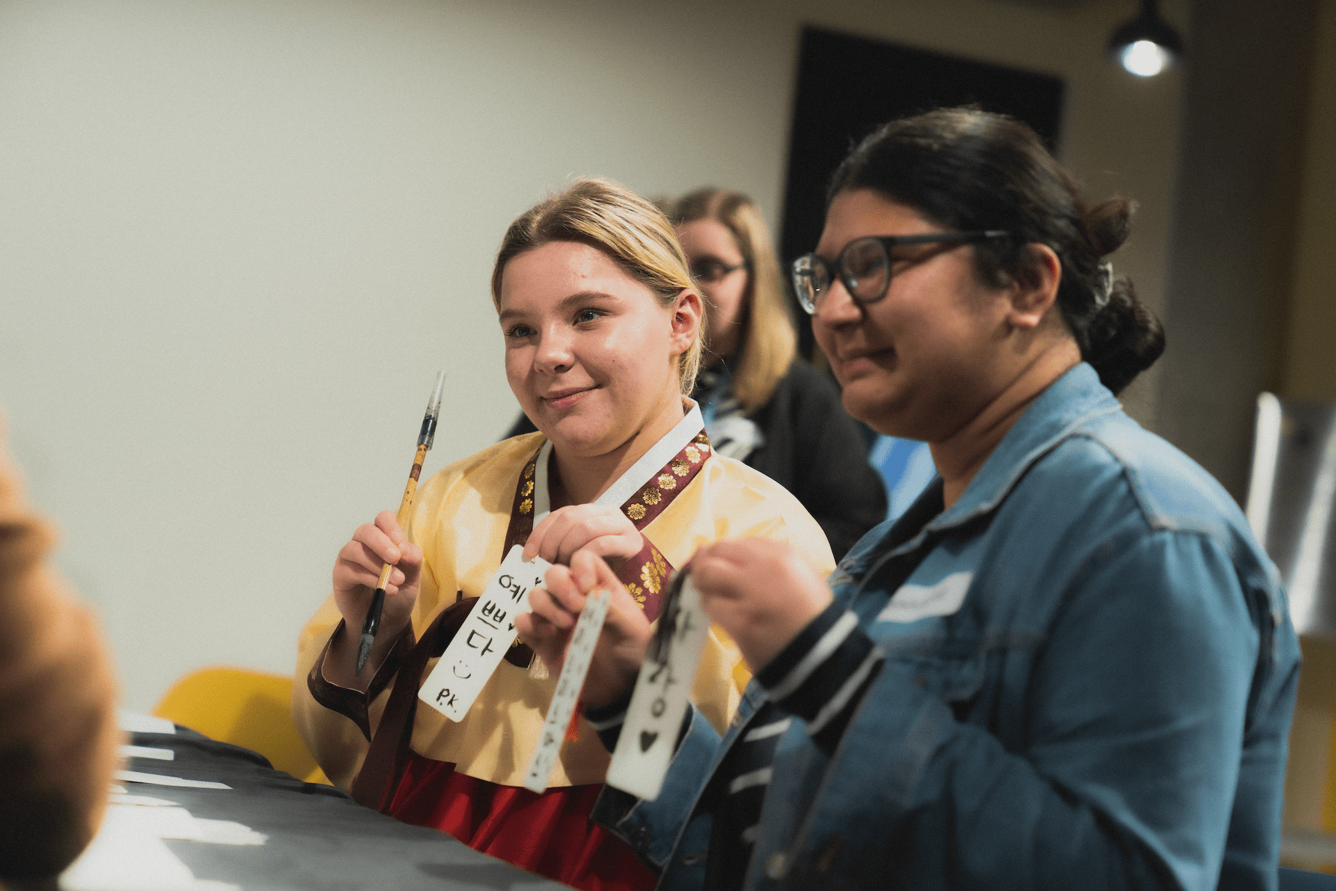People engaging in a traditional Korean calligraphy activity, holding brushes and paper, in an indoor setting with focused expressions.