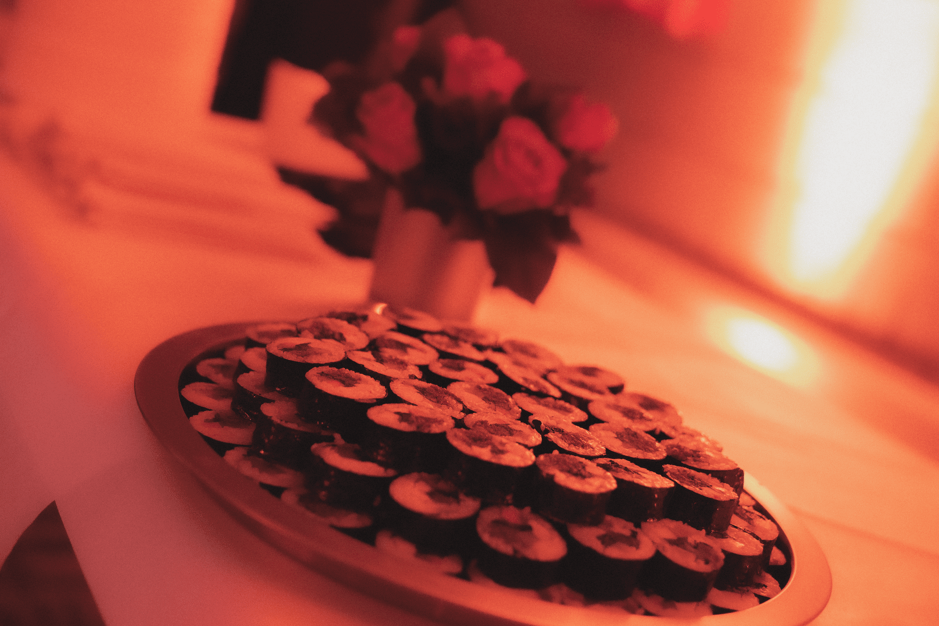 A softly lit room features a tray of sushi rolls and a bouquet of red flowers, creating a warm, inviting atmosphere.