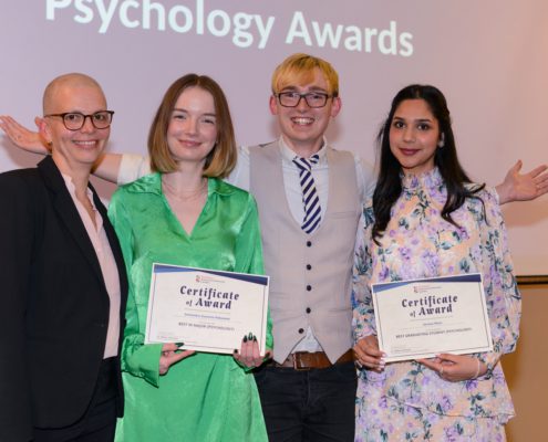 Four people smiling, two holding certificates at a psychology awards event. Casual yet celebratory atmosphere, with a background projection.