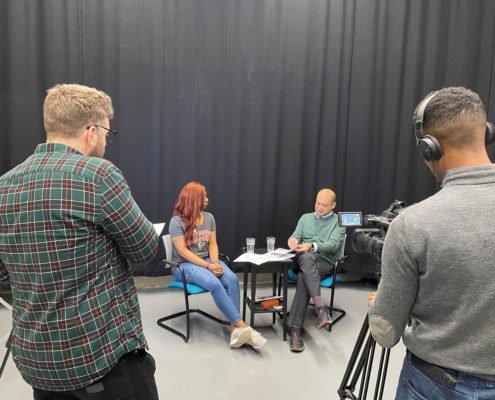 Four people in a studio setting; filming an interview. Two seated near a table, with two crew operating cameras.