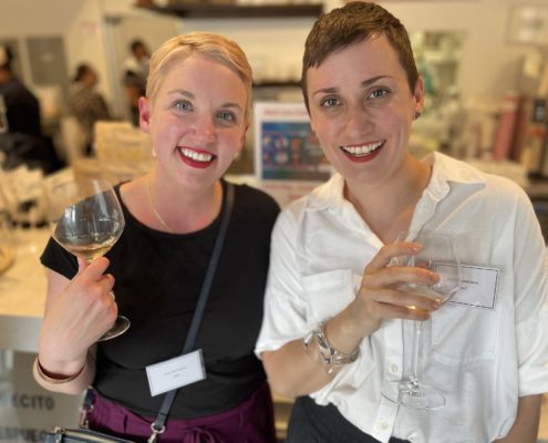 Two people smiling indoors, holding wine glasses. They wear name tags and stand in a lively setting with blurred background elements and other people.