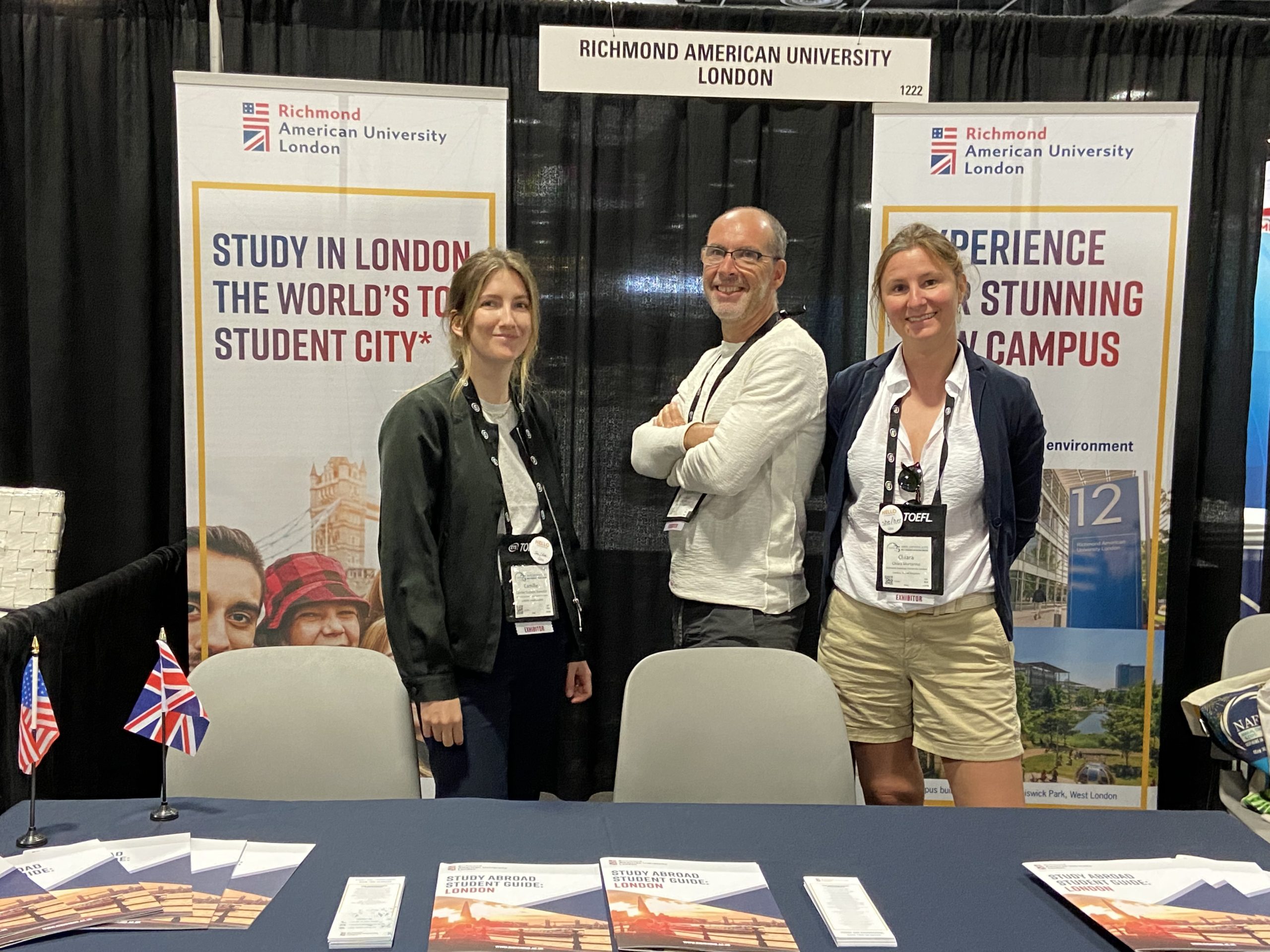 Three people stand at a Richmond American University London booth, promoting study opportunities. Brochures and flags displayed on the table.