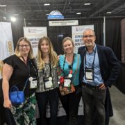 Four people pose at a Richmond American University London booth, with informational banners and various lanyards in a convention setting.