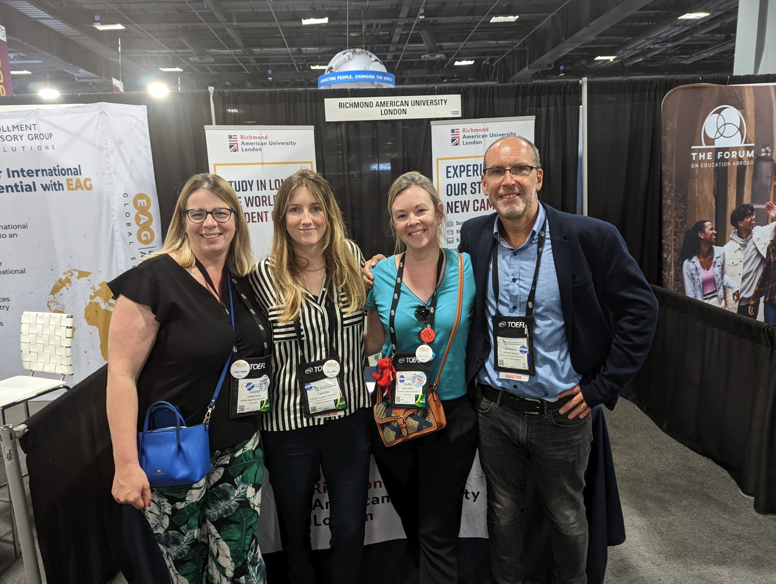Four people pose at a Richmond American University London booth, with informational banners and various lanyards in a convention setting.