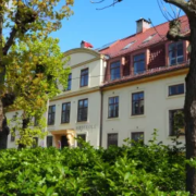 Historic building with red roof amidst lush greenery, trees lining the front. A person is near the entrance labeled "House Sole." Bright, sunny day.
