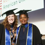 Two people in graduation attire smiling, standing together at a ceremony. A screen in the background displays academic institution logos.