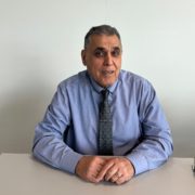 A person in a blue shirt and tie sits at a table against a plain background, looking at the camera.