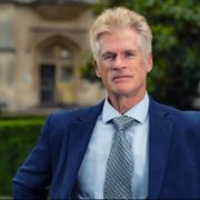 A person in a suit stands confidently outside a historic building with arched entrance and trimmed hedges, surrounded by greenery.
