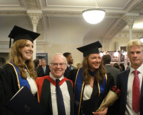 Four people celebrating a graduation, two in caps and gowns, indoors with ornate ceilings, one holding a bouquet. Event appears formal.
