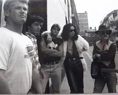 A group of five people stand casually in an urban street setting, with one person holding a guitar. No landmarks are visible.