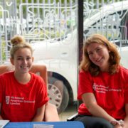 Two people wearing red Richmond American University London shirts sit smiling indoors, next to a window with a decorative pattern and a parked vehicle outside.