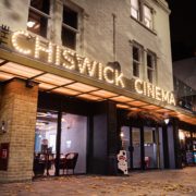 The Chiswick Cinema entrance at night features illuminated signage and inviting interiors, with fallen autumn leaves scattered on the pavement.