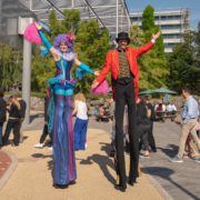 Two performers on stilts entertain a crowd in a plaza, surrounded by greenery and modern architectural structures. Bright costumes add vibrant charm.