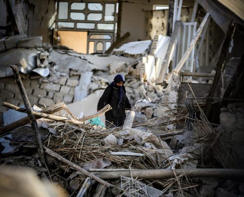 A person walks through rubble and debris of a damaged building, highlighting destruction and aftermath of a disaster. No landmarks identifiable.