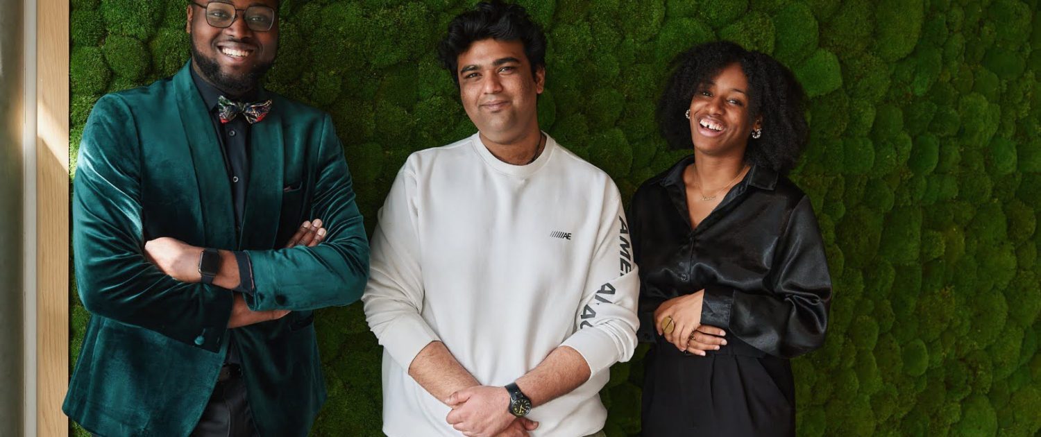 Three people stand smiling against a lush green textured wall, wearing stylish clothing in a well-lit indoor space.