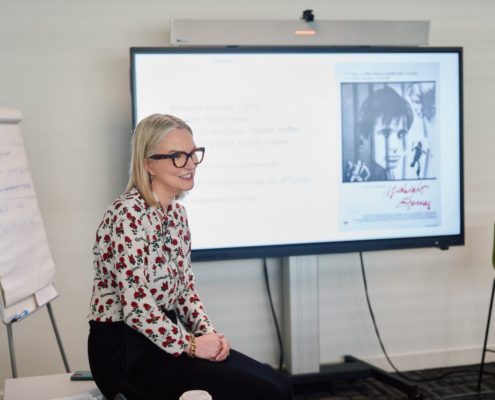 A person with glasses, floral top, sits beside a presentation screen showing a black-and-white image, with a flip chart in the background.
