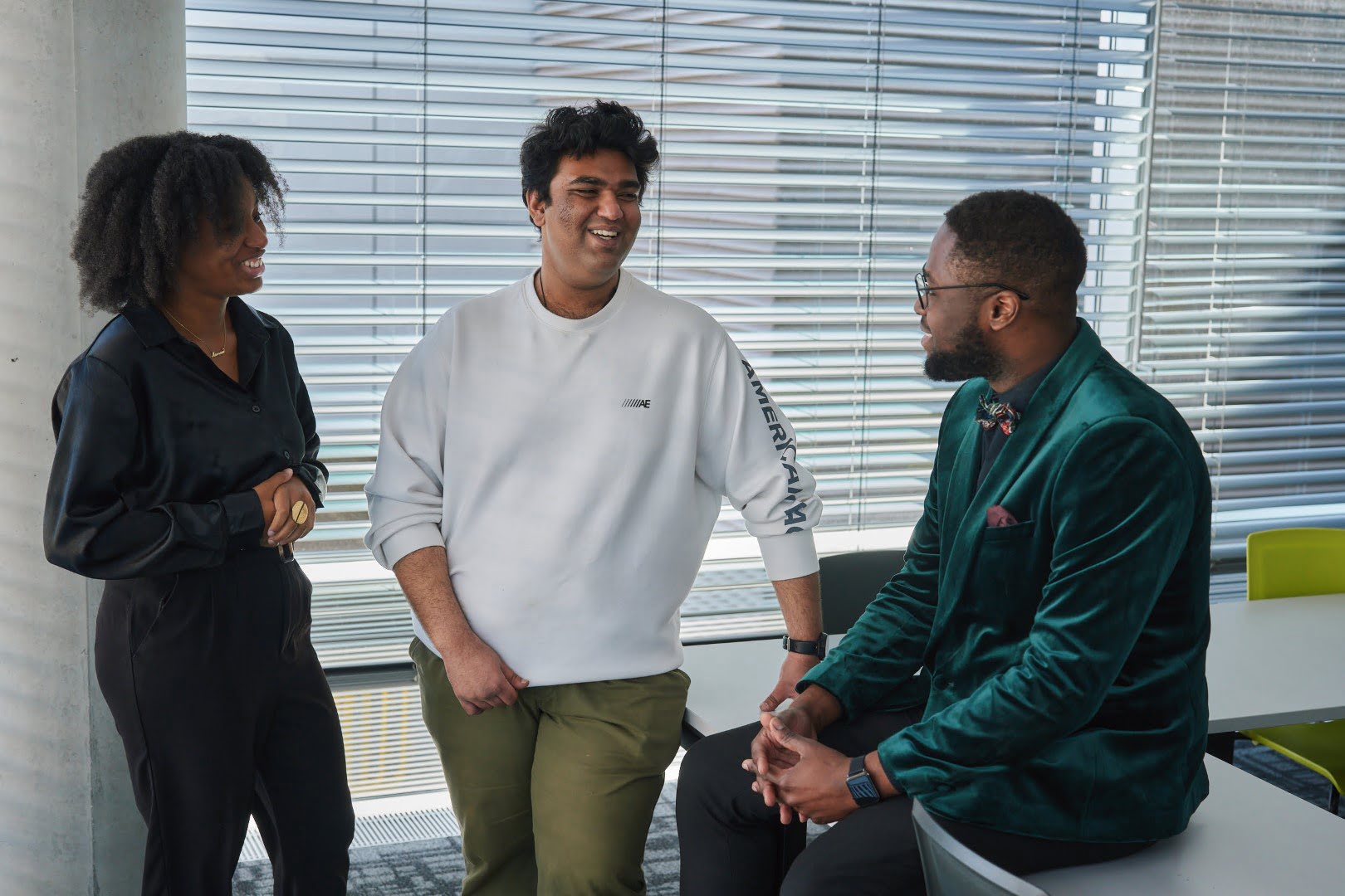 Three people are talking and smiling in a modern office space with blinds, desks, and chairs. Casual and professional attire is visible.
