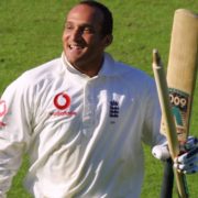 A person in a sports uniform is holding cricket equipment while standing on a grassy outdoor field.