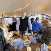 People gather under a lit tent, engaging in conversation, and food sharing. The atmosphere seems casual and convivial, with a focus on interaction and communion.