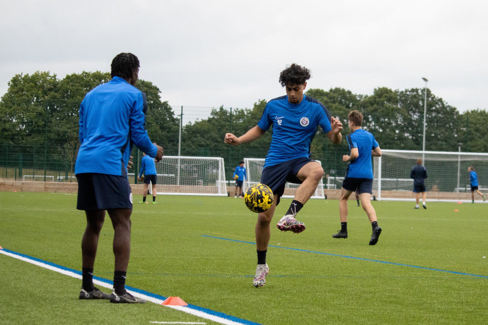 A group of soccer players in blue jerseys are training on a synthetic field. One individual is controlling a yellow and black soccer ball.