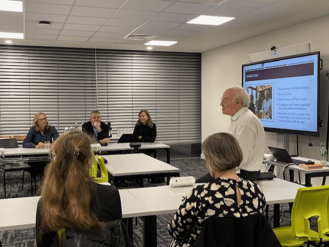 A group of people sits around tables in a well-lit meeting room. One person stands presenting, with a projection screen displaying text behind them.