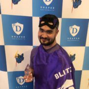 A person in a purple cape and mask holding a trophy, smiling in front of a Draper University step-and-repeat banner.