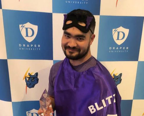 A person in a purple cape and mask holding a trophy, smiling in front of a Draper University step-and-repeat banner.