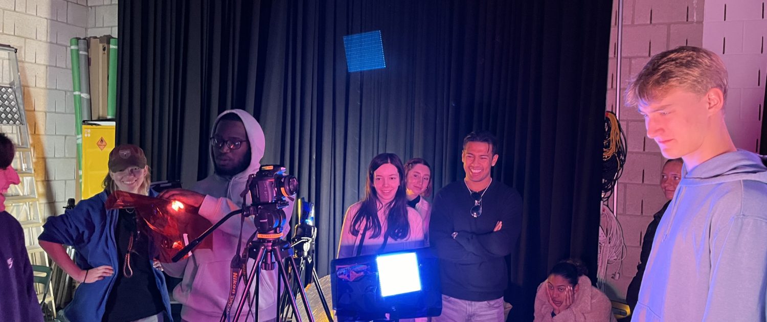 A group of people stands around film production equipment, with cameras, lights, and monitors in an indoor setting, possibly on a set or studio.