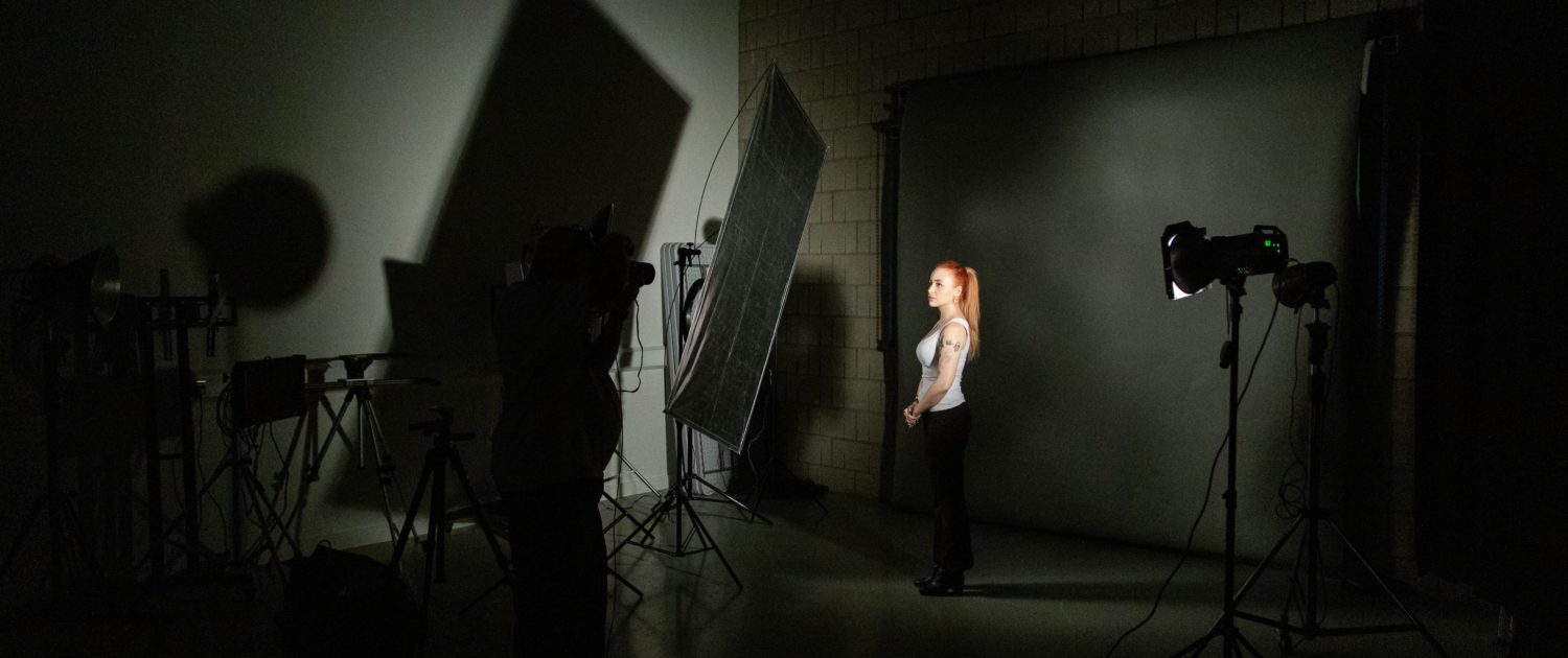 A person is photographing another individual in a dimly lit studio with professional lighting equipment casting sharp shadows on the wall.