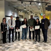 Seven people stand indoors, holding certificates. They seem pleased, possibly at a graduation or recognition event. The space has modern lighting and a coffee bar.