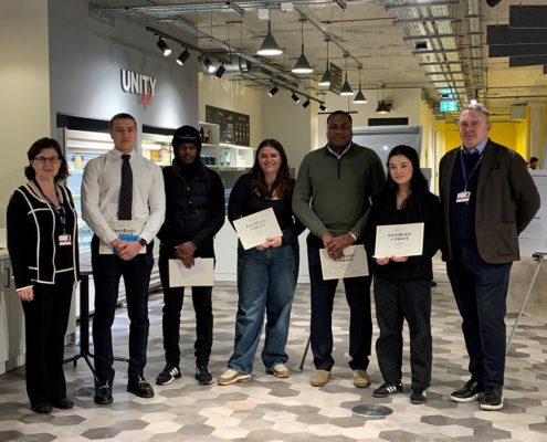 Seven people stand indoors, holding certificates. They seem pleased, possibly at a graduation or recognition event. The space has modern lighting and a coffee bar.