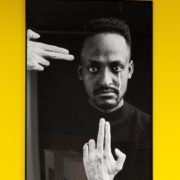 A framed black and white photograph of a person with intense gaze, framed by two hands, against a bright yellow background.