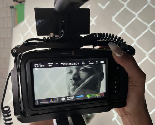 A person's hand is holding a professional video camera with a screen showing a black and white image of a person's face, implied to be the subject being filmed.