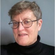 The image shows a person with short hair wearing round glasses and a black turtleneck, against a plain blue background, slightly smiling.