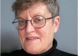 The image shows a person with short hair wearing round glasses and a black turtleneck, against a plain blue background, slightly smiling.