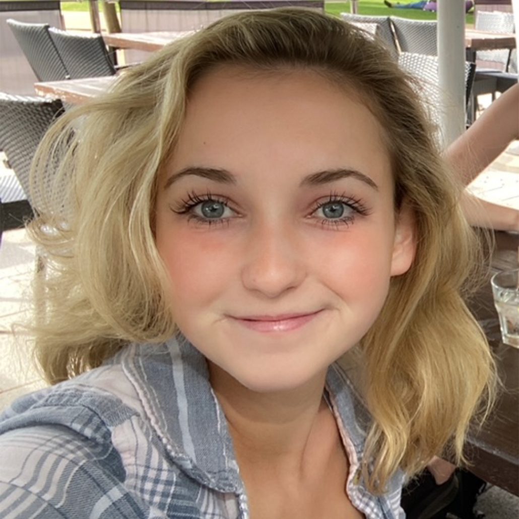 A smiling person with blonde hair is pictured, featuring clear blue eyes, in a casual plaid shirt, outdoors with seating in the background.