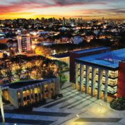 An urban landscape at dusk with illuminated buildings, a modern structure in the foreground, and a vibrant sunset sky blending into a city silhouette.