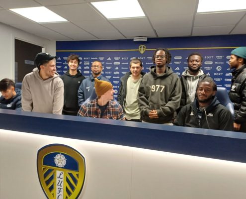 A group of people smiling in front of a counter with a Leeds United Football Club logo displayed. They appear to be in a press room.