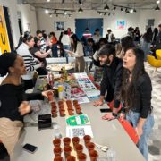 Several people are gathered in a modern, well-lit room with artwork, engaged in conversations and activities around tables with plants and food.