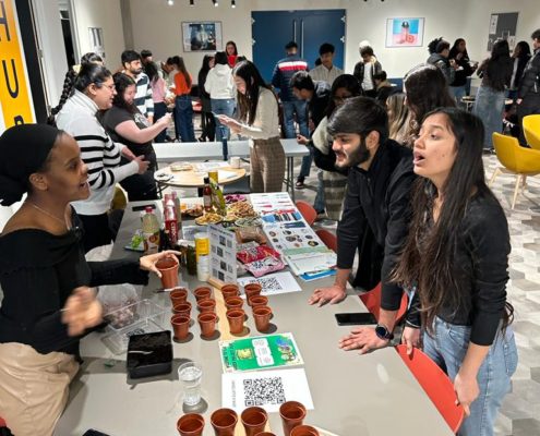 Several people are gathered in a modern, well-lit room with artwork, engaged in conversations and activities around tables with plants and food.