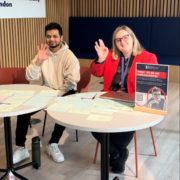 Two people are sitting at a table with papers, smiling and making peace signs, next to a sign about becoming an ambassador.