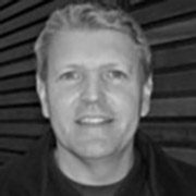 The image shows a grayscale portrait of a smiling person with short hair, standing in front of a striped background. The person wears a dark top.